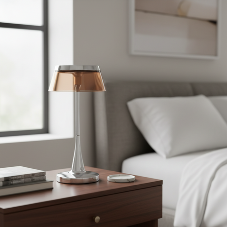 A modern table lamp with a brushed silver metal stem and base, and a translucent copper-colored shade, sitting on a dark wood bedside table in a softly lit bedroom.