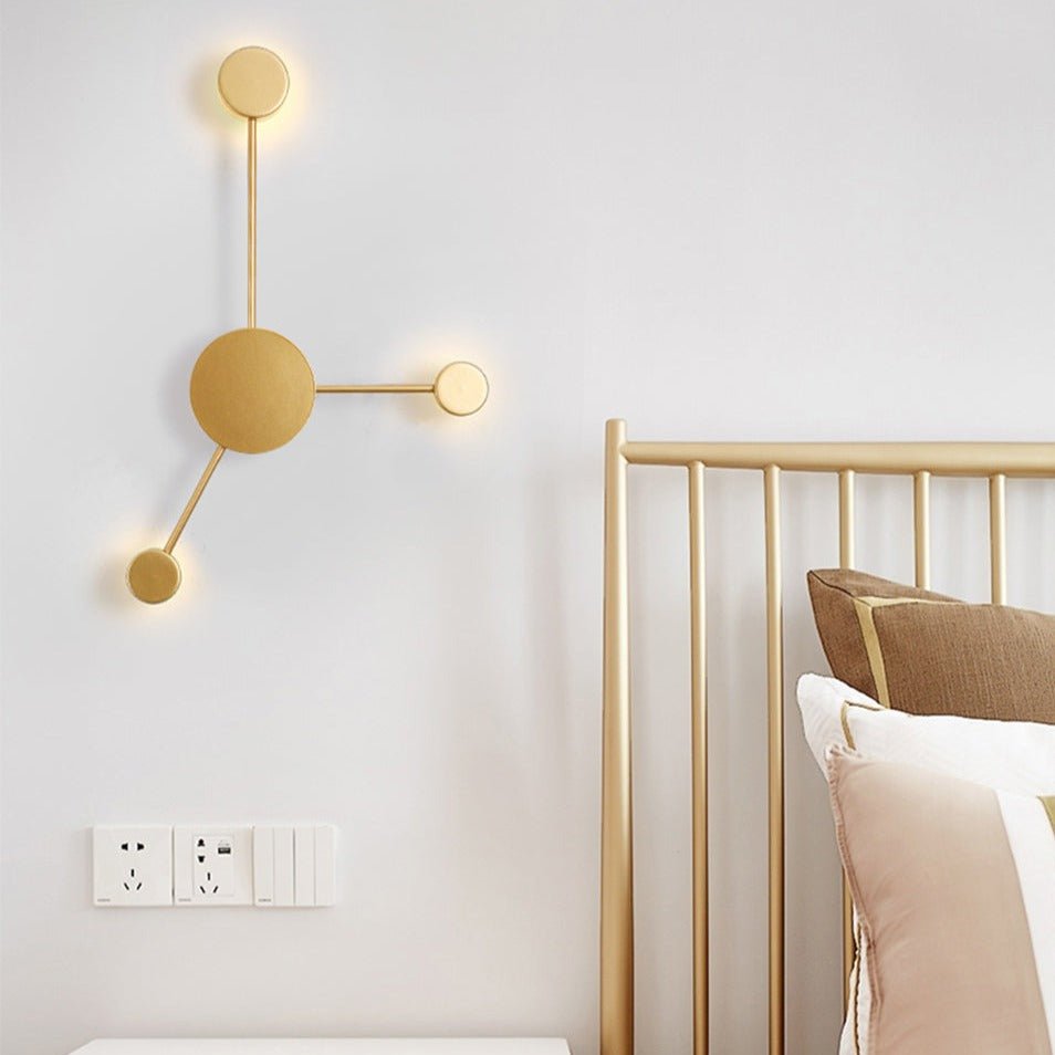 Gold three head wall sconce lit on a white wall beside a brass bed