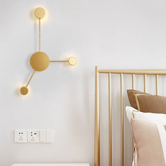Gold three head wall sconce lit on a white wall beside a brass bed