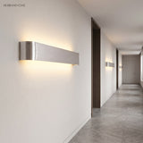 Silver linear wall sconce lit on a white corridor wall