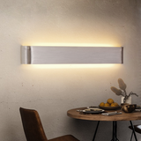 Silver linear wall sconce lit on a plaster wall above a table