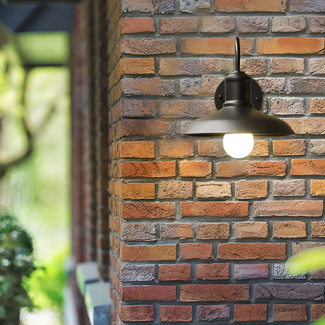 Gooseneck barn light lit on brick beside a climbing plant