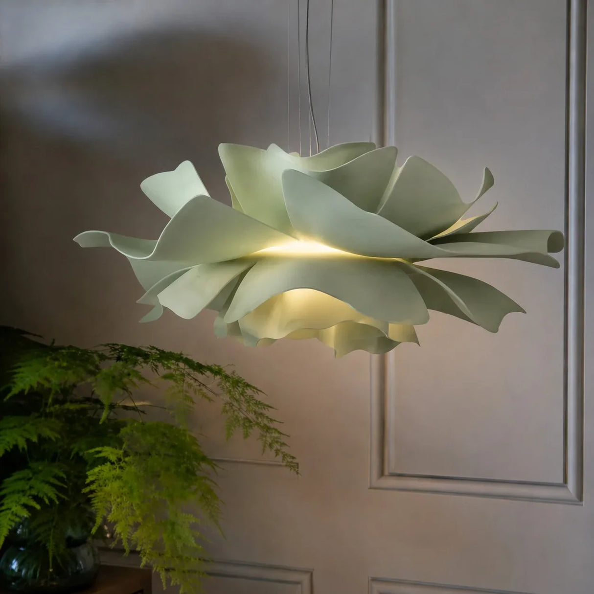 Sage green petal pendant light lit by a panelled wall and fern