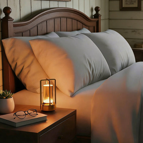 Gold cordless lamp lit on a nightstand beside pillows and reading glasses