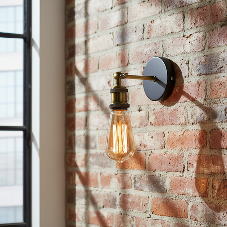 Industrial-style wall sconce with a round black metal wall mount, a brass-finished arm, and a clear glass Edison bulb, shown mounted on a red brick wall next to a window.