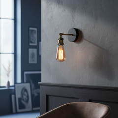 A wall-mounted industrial-style light fixture with a warm glowing Edison bulb, mounted on a textured gray wall above a dark wood headboard in a bedroom setting.