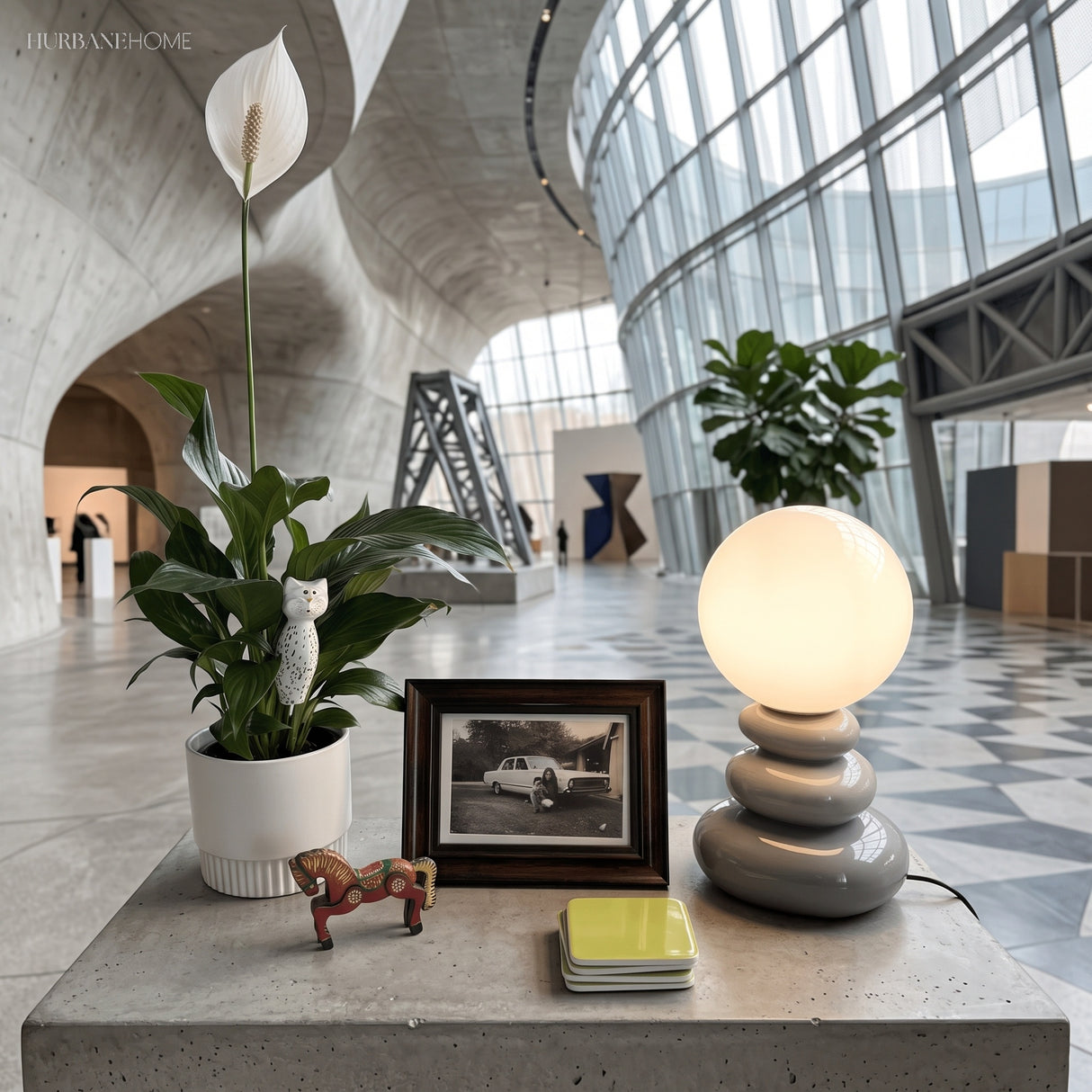 Grey stacked ceramic lamp lit on a concrete ledge beside plants and a frame