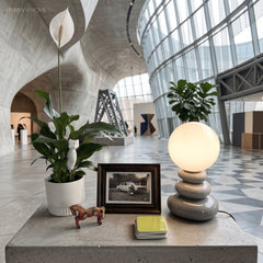 Grey stacked ceramic lamp lit on a concrete ledge beside plants and a frame