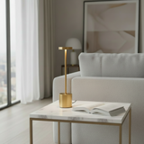 A modern gold table lamp with a cylindrical base and rectangular shade sits on a white marble-top side table in a bright, contemporary living room next to a light gray sofa.