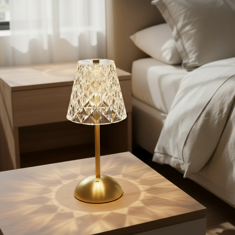 A table lamp with a polished gold-finished metal base and stem, featuring a tapered crystal or faceted glass shade that glows warm white when lit, shown against a plain white background.