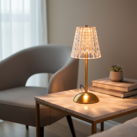 A table lamp with a warm brass-finished metal base and stem, featuring a pleated, faceted clear glass shade that glows with a warm light, set against a plain white background.