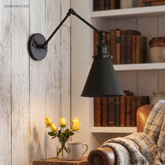 Black swing arm sconce lit warm beside books and a leather chair