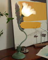 Tall flower table lamp lit on a sideboard with foliage and framed art behind