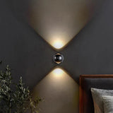 Black orb sconce lit on a grey wall beside a bed and a potted olive tree