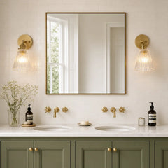Two brass sconces either side of a mirror above green bathroom cabinets