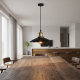 A modern black pendant light with a brass-toned collar and glowing bulb, hanging above a wooden dining table in a bright, contemporary kitchen and dining room.