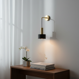 A modern wall-mounted sconce with a black cylindrical shade and a gold-finished arm and wall mount, shown lit on a white wall above a wooden console table with books and a small plant.