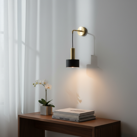 A modern wall-mounted sconce with a black cylindrical shade and a gold-finished arm and wall mount, shown lit on a white wall above a wooden console table with books and a small plant.