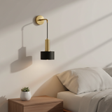 A modern wall-mounted lamp with a gold wall plate and cylindrical gold and black shades, shown lit in a bedroom setting next to a bed and wooden bedside table.