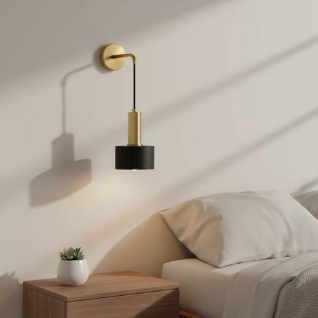 A modern wall-mounted lamp with a gold wall plate and cylindrical gold and black shades, shown lit in a bedroom setting next to a bed and wooden bedside table.