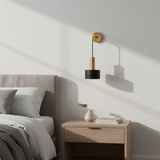 A modern wall-mounted sconce with a gold and black finish, mounted on a white wall above a light wood nightstand in a minimalist bedroom setting.