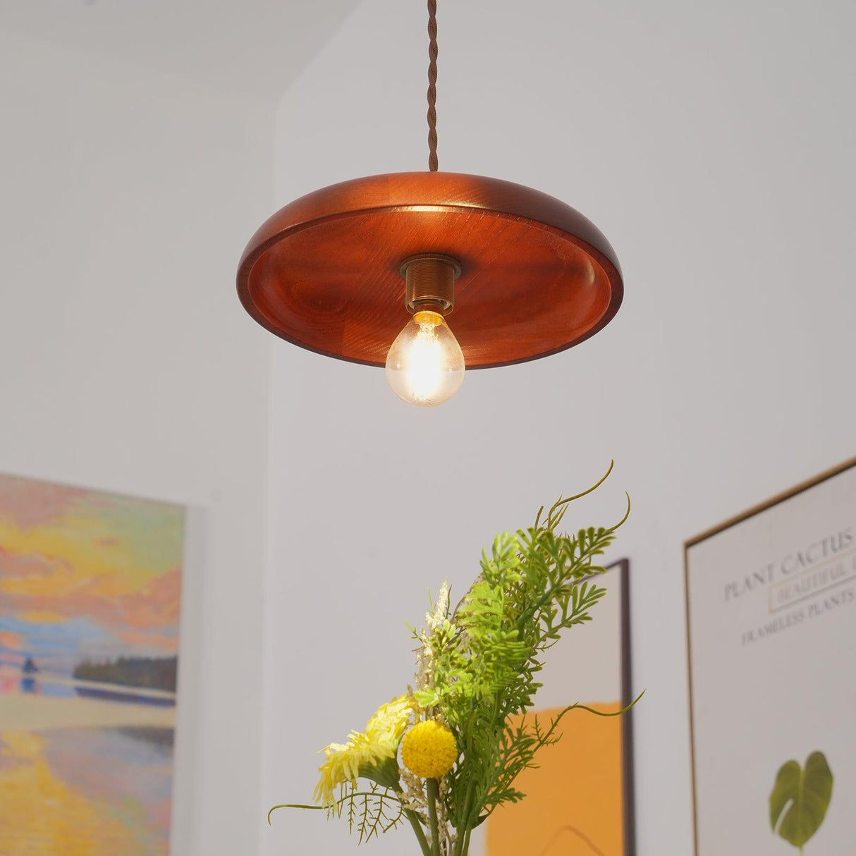 Wood pendant lit in a white hallway above framed prints and a vase of foliage