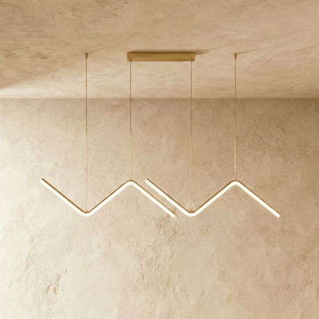 Row of gold zigzag pendant bars lit against a pale textured plaster wall